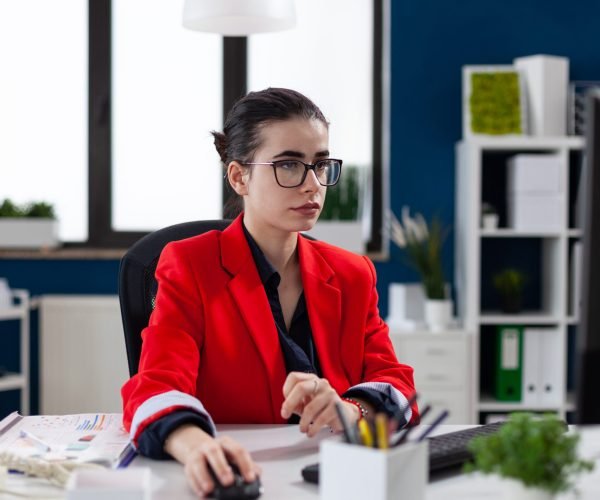 Focused businesswoman in corporate office workplace, working on computer, making financial analysis. Concentrated entrepreneur using mouse with wireles technology sitting at desk.